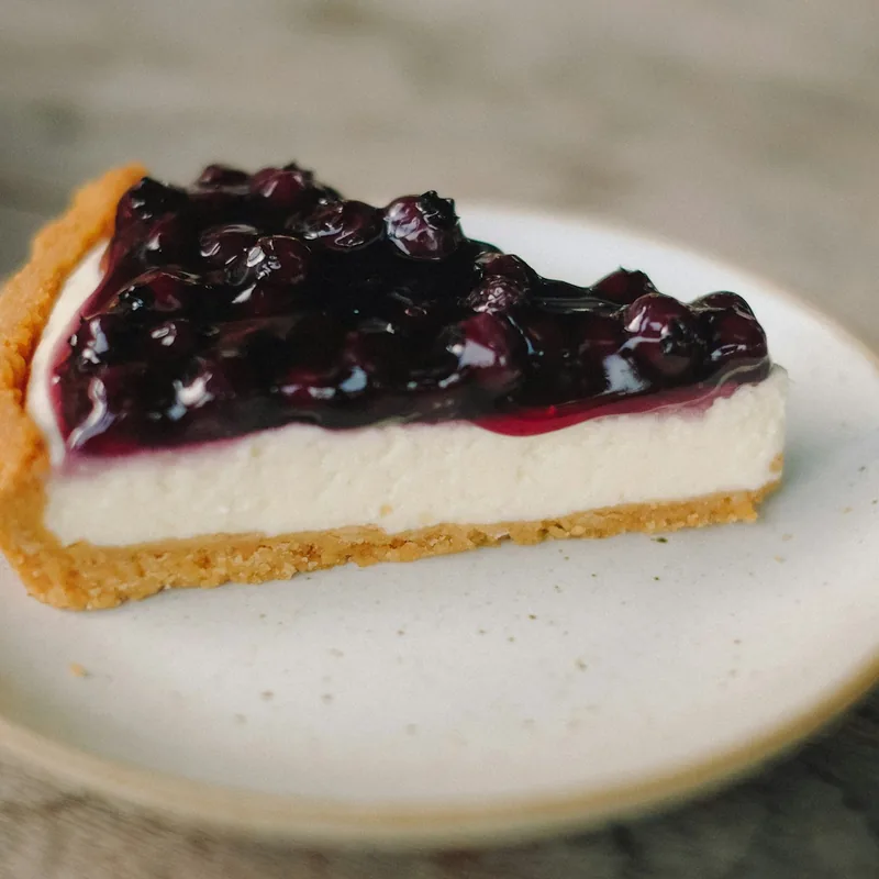 En bit cheesecake med blåbärstopp på vit tallrik